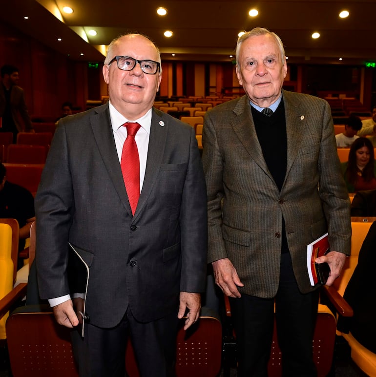 Dos hombres sonrientes, uno con traje gris y corbata roja, el otro con chaleco marrón y camisa negra, posan en un auditorio.