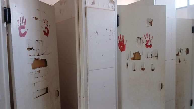 Tres puertas de un baño en mal estado, con huellas rojas y signos de abandono, en un ambiente deteriorado.