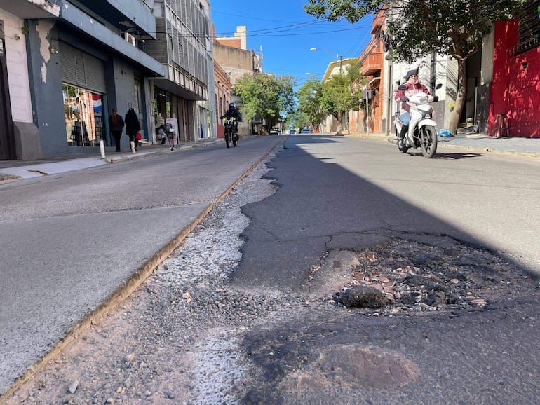 Dos personas caminan por la acera, un ciclista pasa por la calle junto a un motociclista, con un bache visible en la calzada.