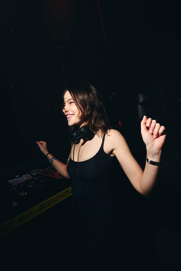 Anfisa Letyago sonríe mientras actúa como DJ en un ambiente oscuro, con luces de fondo y una mesa de mezcla frente a ella.