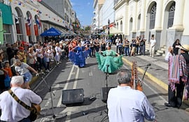 Dos mujeres bailan, una con vestido azul y otra con vestido verde, rodeadas de un público aplaudiendo en un ambiente festivo.