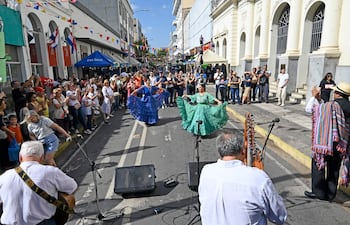 Dos mujeres bailan, una con vestido azul y otra con vestido verde, rodeadas de un público aplaudiendo en un ambiente festivo.