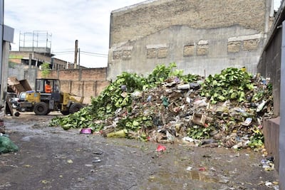 Operador con overol naranja maneja cargadora amarilla en mercado lleno de basura y desechos, bajo un ambiente desordenado.