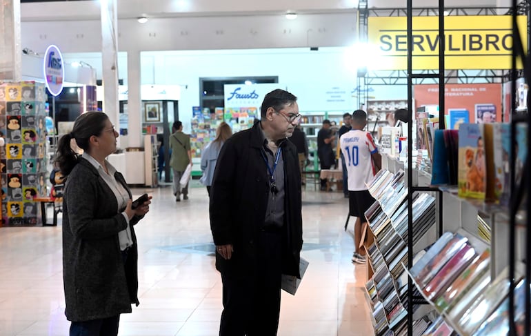 Mujer con abrigo gris y hombre de cabello canoso observan libros en un ambiente iluminado de la feria.