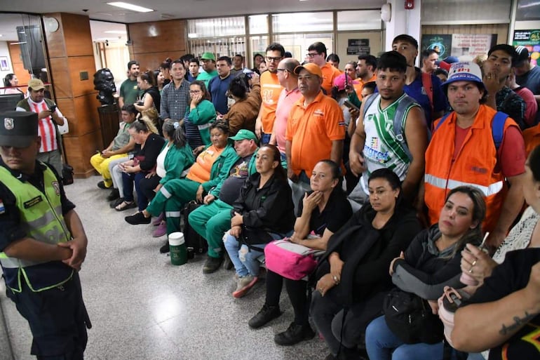 Oficial de seguridad en uniforme observa a un grupo diverso de personas en sala gubernamental. Ambiente de espera y conversaciones.