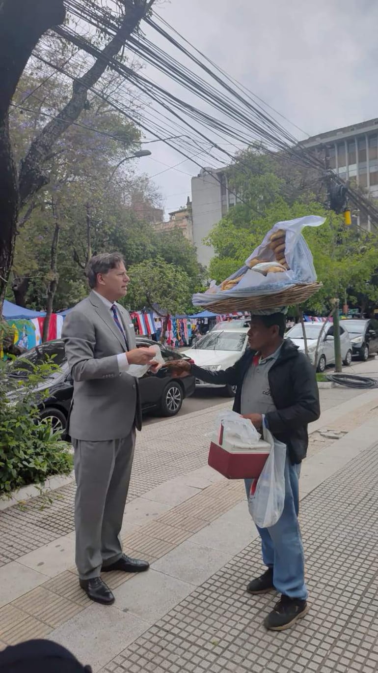 Christopher Landau, vestido con traje gris, dialoga con un vendedor que sostiene una canasta de comida en la acera.