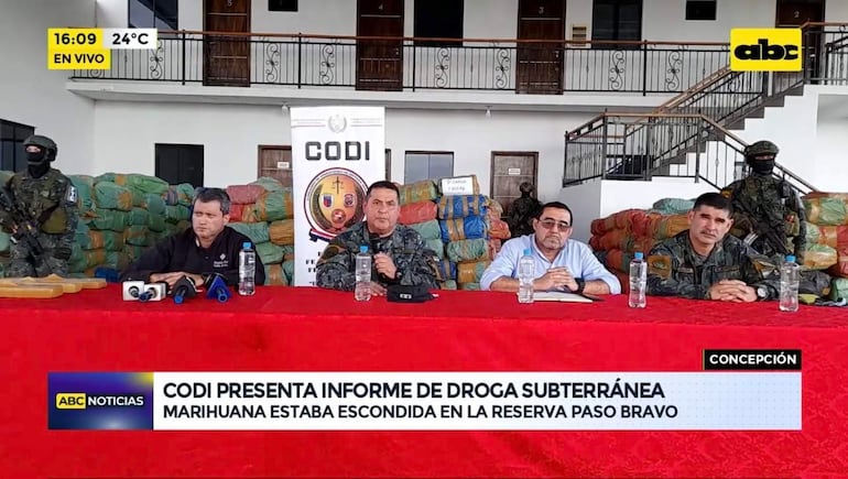 Hombres en uniforme militar y civil durante conferencia, con expresiones de atención y un ambiente formal decorado.