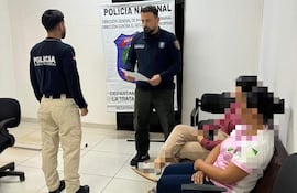Oficial de policía de pie con documento, mientras dos civiles observan atentamente en una oficina con cartel de Policía Nacional.