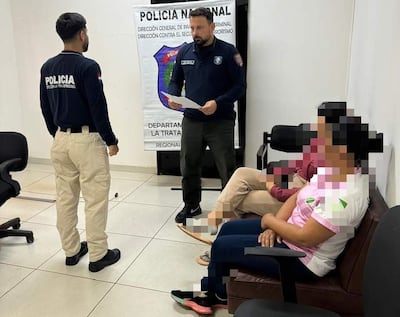 Oficial de policía de pie con documento, mientras dos civiles observan atentamente en una oficina con cartel de Policía Nacional.