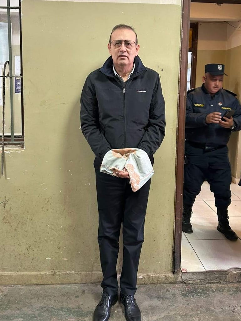 Erico Galeano de pie, vestido con abrigo oscuro y pantalones negros, en un ambiente administrativo junto a un oficial de policía.