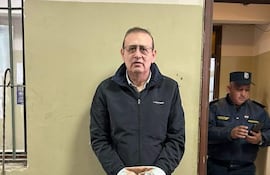 Erico Galeano de pie, vestido con abrigo oscuro y pantalones negros, en un ambiente administrativo junto a un oficial de policía.