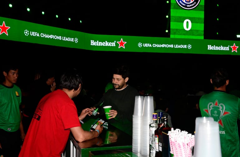 Hombre con camiseta negra y vaso verde interactúa con otro hombre de camiseta roja en evento iluminado con anuncios de Heineken.