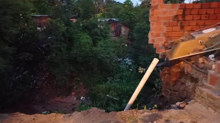 Terreno en deslizamiento con muro de ladrillos derrumbado rodeado de vegetación densa y residuos visibles.