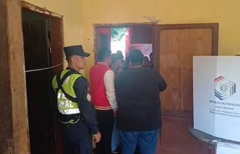 Grupo de personas en sala de votación, con oficial de seguridad y cabina electoral. Ambiente tranquilo, paredes amarillas.