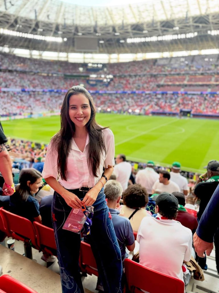 Mujer de cabello largo y lacio, con blusa blanca y pantalones de mezclilla, sonríe en un estadio lleno de público.