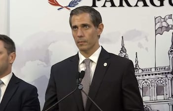 Marco Riquelme en traje oscuro y corbata clara, hablando con seriedad mientras otro hombre escucha en un ambiente formal.