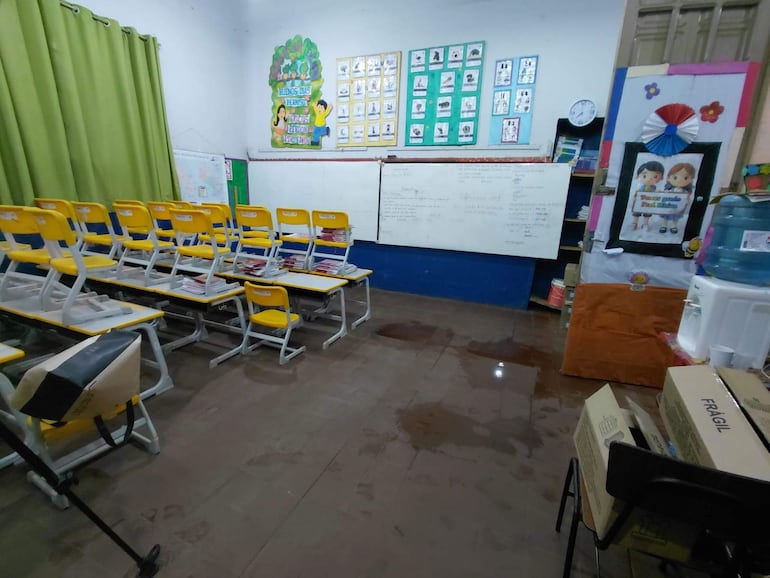 Aula vacía con escritorios y sillas amarillas apiladas, marcas de agua en el suelo y carteleras en las paredes.