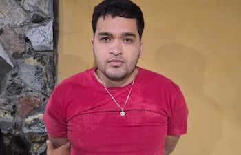Sergio Francia Bernal, con camiseta roja y pantalones oscuros, de pie con expresión seria mientras un agente lo detiene.