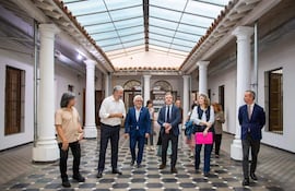 Grupo de seis personas en traje, conversando en pasillo con columnas blancas y luz natural de techo de vidrio.