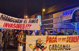 Grupo de hinchas con camisetas amarillas y negras sosteniendo pancartas contra el bingo, en un ambiente nocturno.