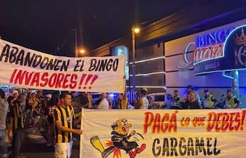 Grupo de hinchas con camisetas amarillas y negras sosteniendo pancartas contra el bingo, en un ambiente nocturno.
