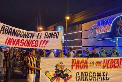 Grupo de hinchas con camisetas amarillas y negras sosteniendo pancartas contra el bingo, en un ambiente nocturno.