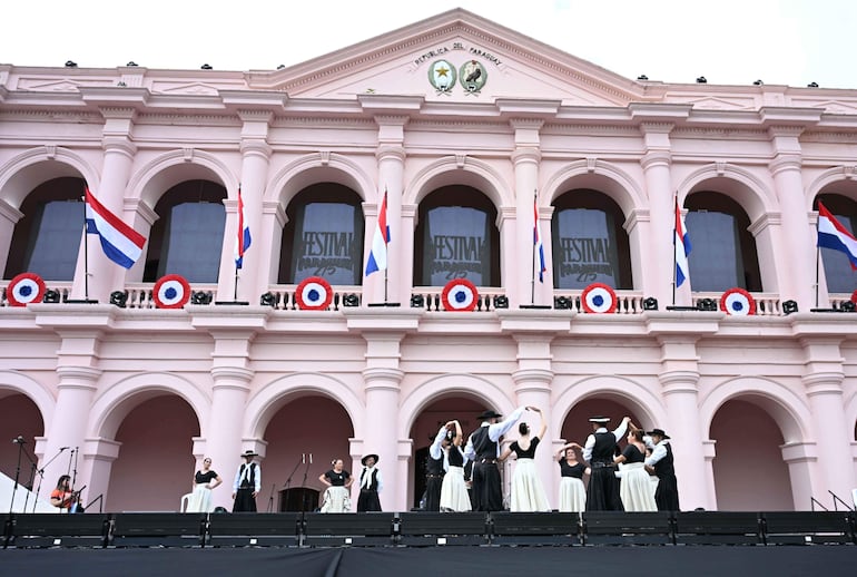 Bailarines en trajes tradicionales negros y blancos, danzando frente a un escenario adornado con banderas, en un ambiente festivo.