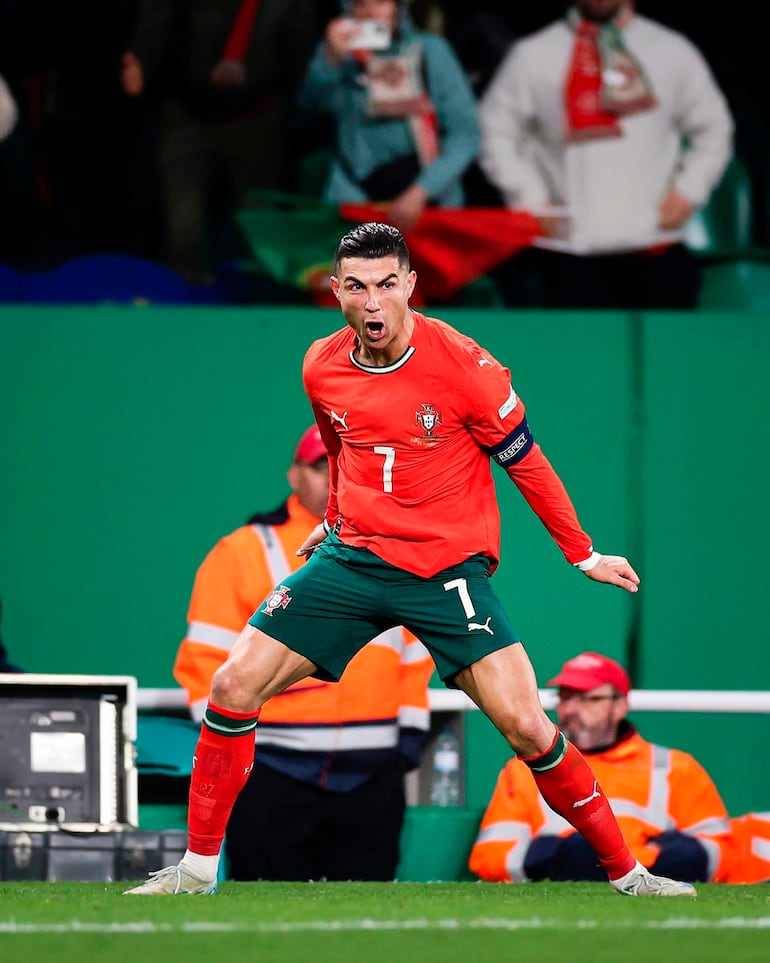 Cristiano Ronaldo en camiseta naranja y pantalones verdes, celebrando un gol con expresión de euforia y afición animando detrás.