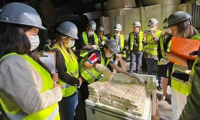 Diez trabajadores en casco y chaleco reflectante observan a una mujer revisando un paquete de dinero en una mesa.