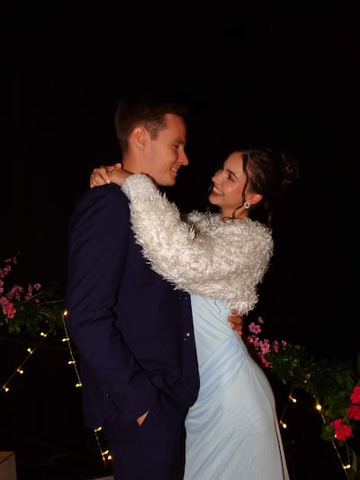 Joshua Duerksen con traje oscuro y Anneke Rahn en vestido azul claro y abrigo blanco, sonriendo en un ambiente festivo con flores.