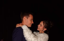 Joshua Duerksen con traje oscuro y Anneke Rahn en vestido azul claro y abrigo blanco, sonriendo en un ambiente festivo con flores.