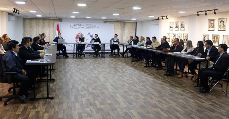 Diversos participantes en vestimenta formal discuten en torno a una mesa. Panel del Ministerio de Educación y Ciencias al fondo.