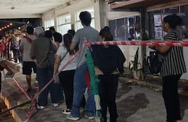 Seis personas en fila con ropa casual, esperando atención médica, en un ambiente urbano con luces de comercio cercano.