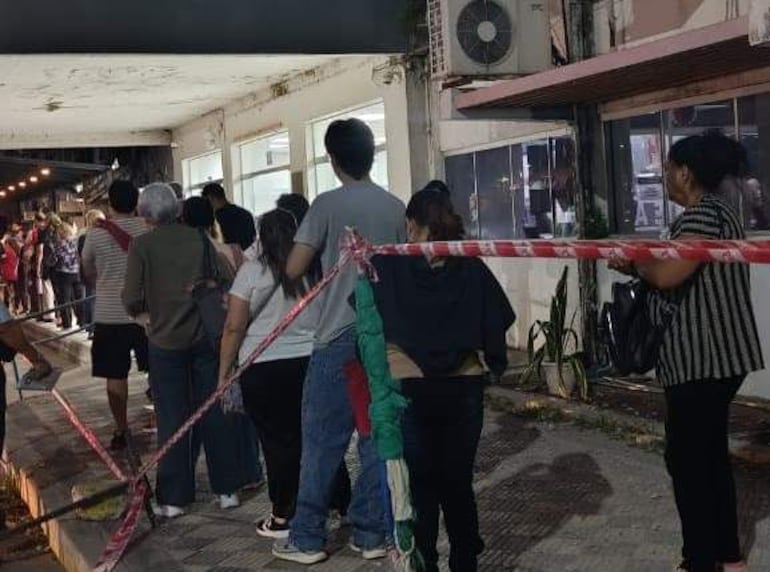 Seis personas en fila con ropa casual, esperando atención médica, en un ambiente urbano con luces de comercio cercano.