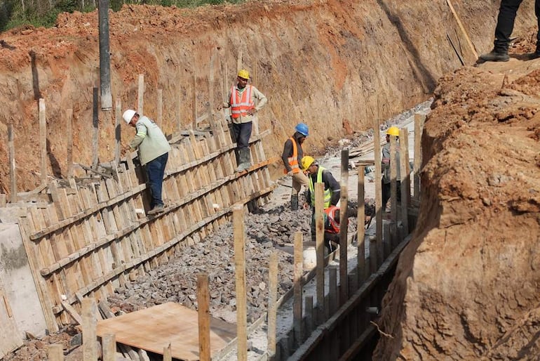 Seis trabajadores en casco en construcción, uno usa herramienta, otros observan. Terreno desparejo y materiales visibles.
