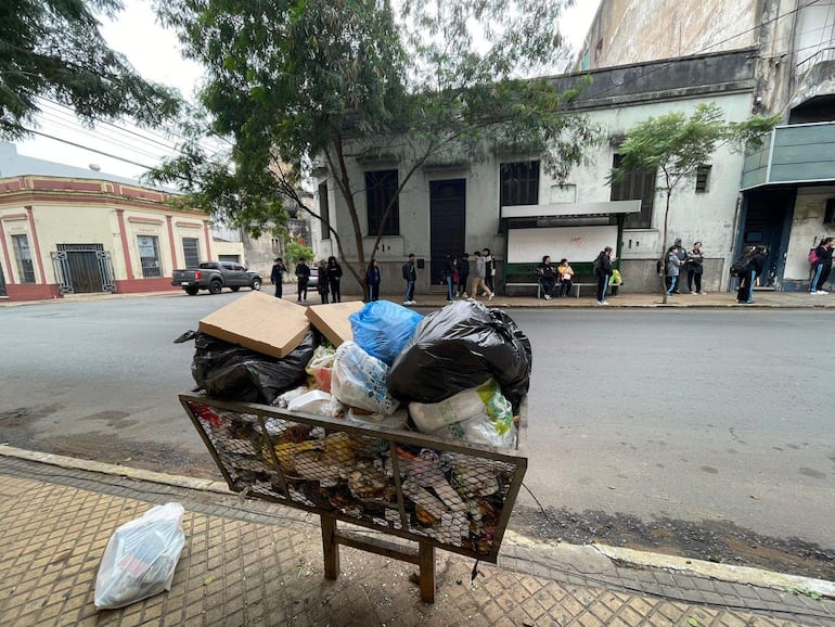 Grupo de jóvenes vestidos de oscuro espera en la acera, con un contenedor de basura repleto en primer plano.