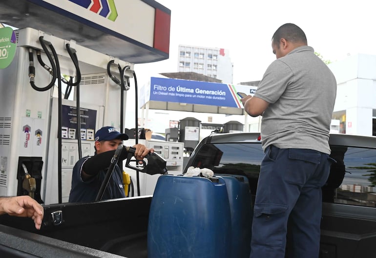 Hombre con gorra y camiseta oscura carga combustible en recipiente azul, mientras otro hombre con camiseta gris usa su teléfono móvil.