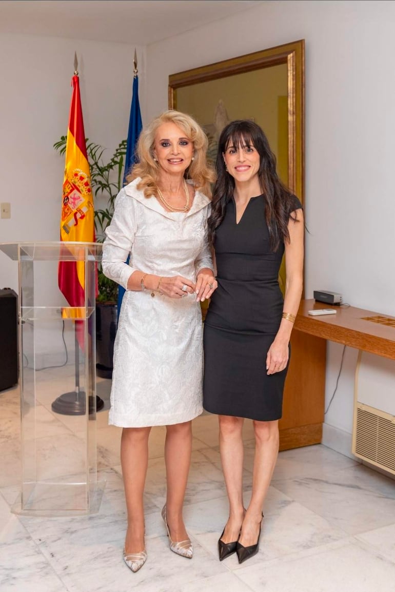 Dos mujeres posando: una en vestido blanco y otra en negro, en un ambiente elegante con banderas de España en el fondo.