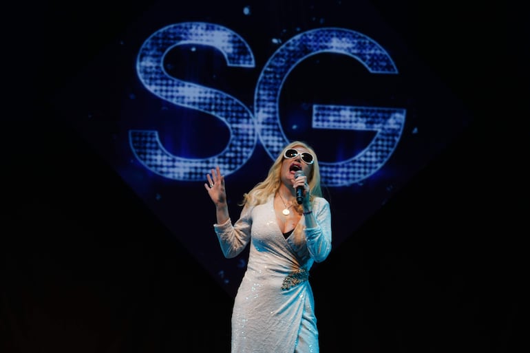 Fátima Flórez en escenario con vestido blanco brillante y gafas de sol, sosteniendo un micrófono mientras interpreta.