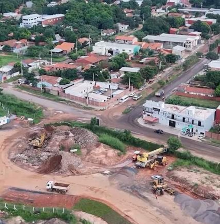 Vista aérea de obra en construcción con maquinaria, camiones y área urbana mezclada con edificios y viviendas, sin personas visibles.