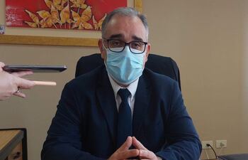 Humberto Otazú sentado en un escritorio con traje oscuro y mascarilla, manos juntas, en un ambiente de oficina profesional.
