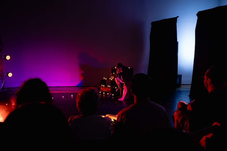 Grupo de personas observando un performance en un escenario iluminado, con luces rosa y azul y ambiente íntimo.
