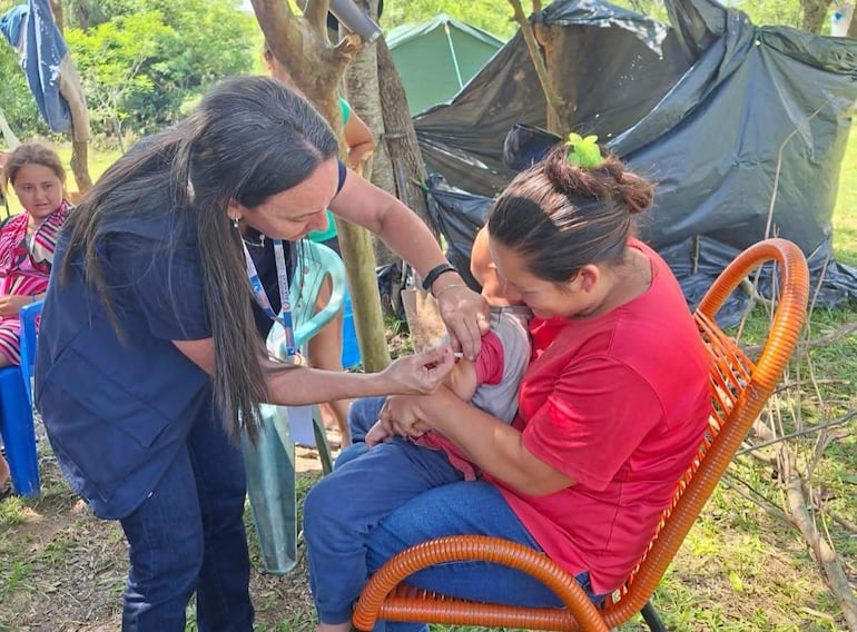 Mujer en camiseta oscura vacuna a niño en piernas de su madre, quien lleva camiseta roja, en área de refugio al aire libre.