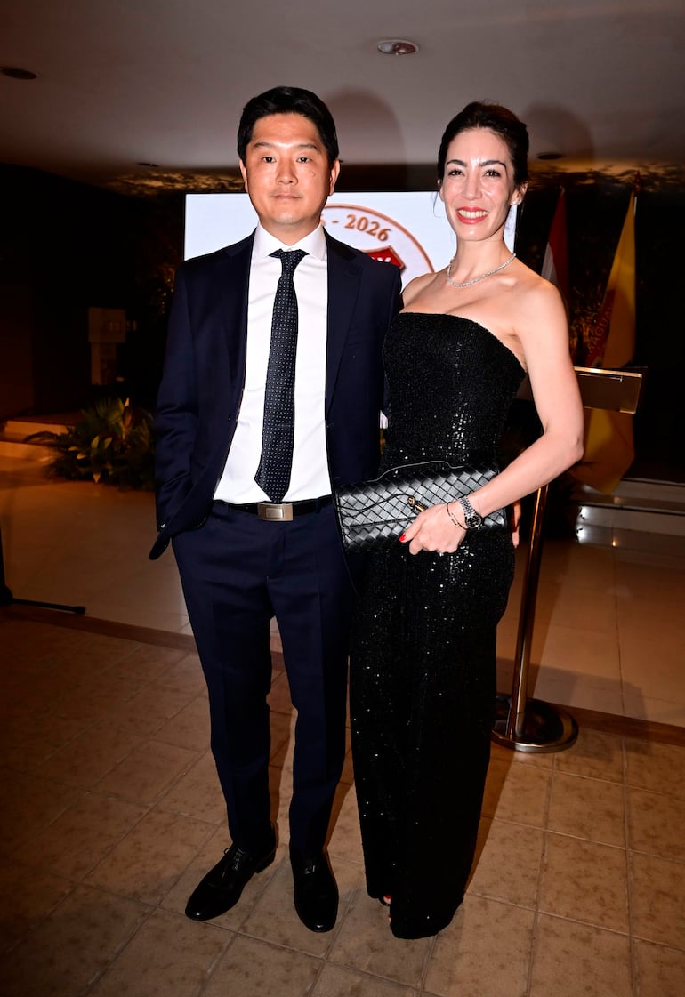 Hombre con traje oscuro y camisa blanca junto a mujer en vestido negro, ambos sonrientes en un entorno elegante.