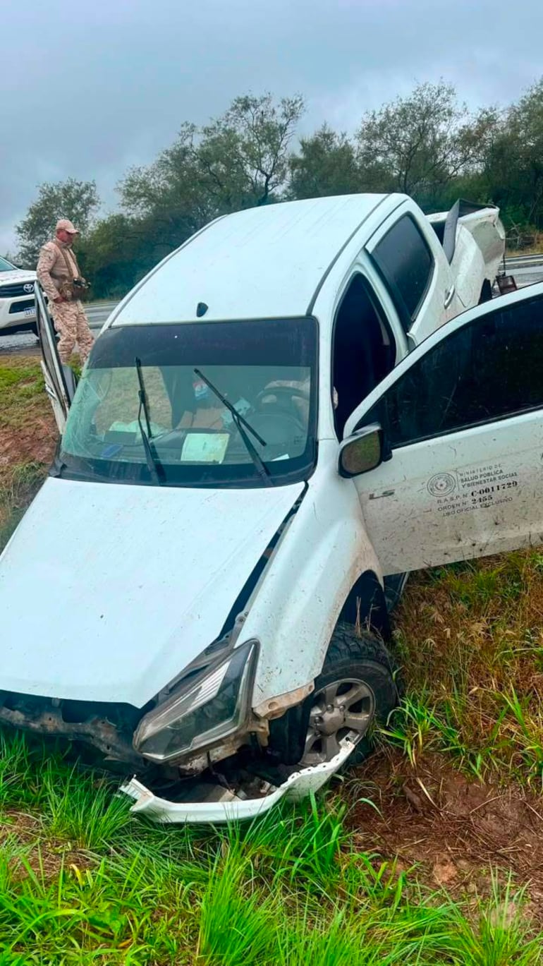 Hombre con uniforme observa una camioneta blanca accidentada con parachoques colgando en un terreno grassoso bajo cielo nublado.