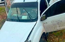 Hombre con uniforme observa una camioneta blanca accidentada con parachoques colgando en un terreno grassoso bajo cielo nublado.