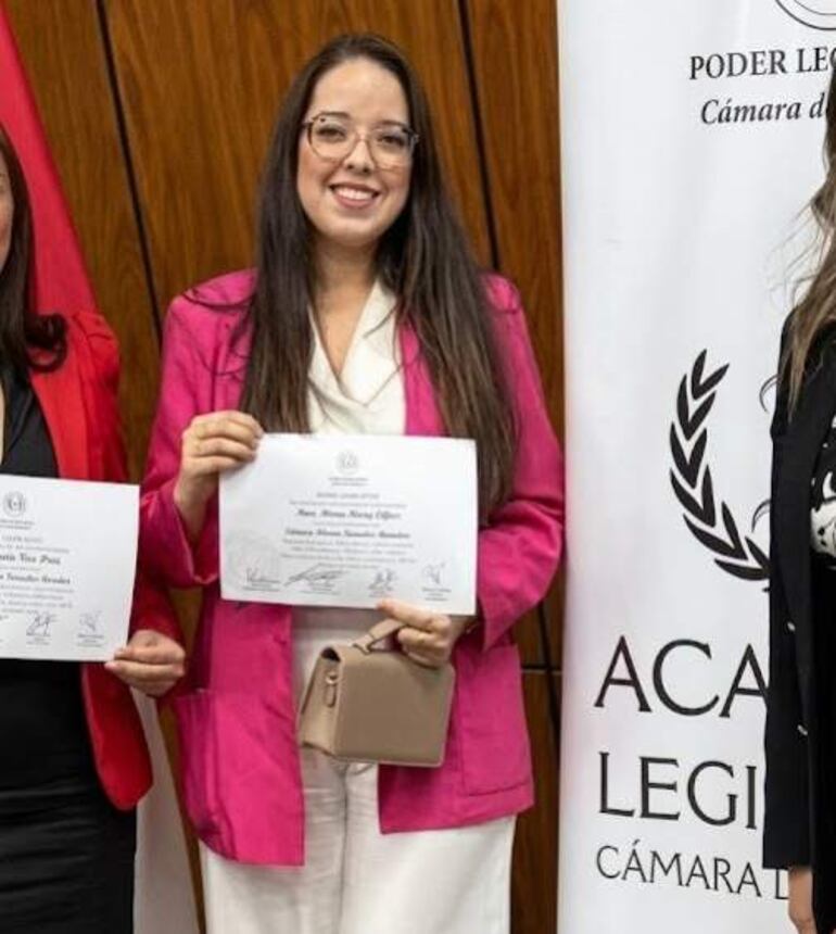 Susana Adriana Sánchez Delgado sonríe con blazer rosa, recibiendo un certificado junto a dos mujeres con atuendos rojo y negro.