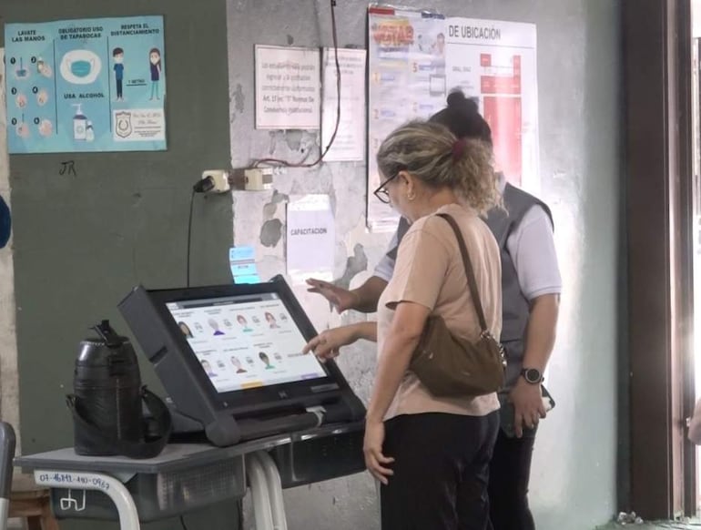 Mujer de cabello rizado claro, en camiseta clara, interactúa con pantalla, asistida por mujer con chaqueta gris en un centro de votación.