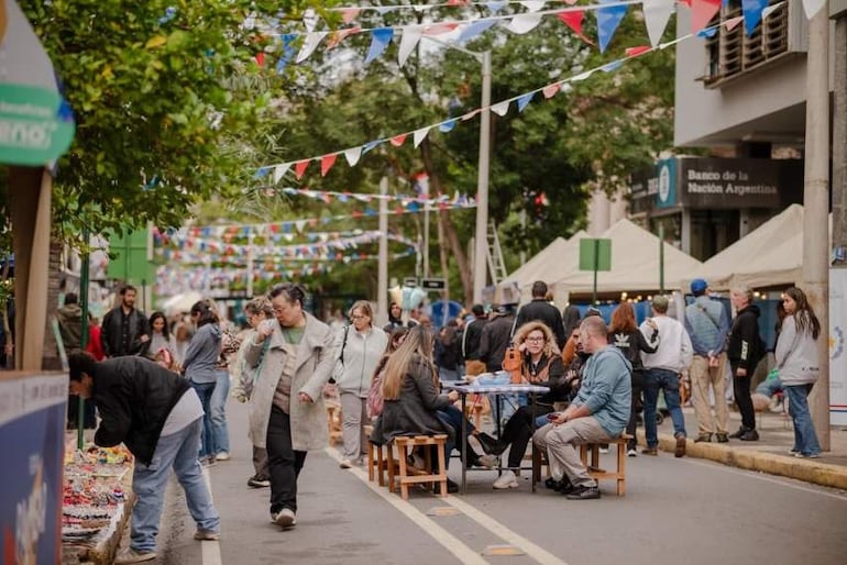 Grupo de personas sentadas y socializando en feria, un hombre de pie con cabello oscuro observa el mercado cercano.