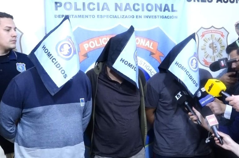 Hombres encapuchados con la palabra 'HOMICIDIOS', rodeados de policías y micrófonos en conferencia de prensa sobre homicidios.